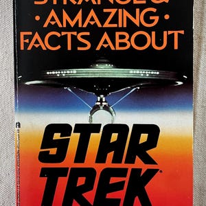 Puede incluir: Un libro de bolsillo titulado "STAR TREK" con el texto "STRANGE & AMAZING FACTS ABOUT" en naranja. La portada presenta una ilustración de nave espacial sobre un fondo degradado de azul, naranja y rojo.