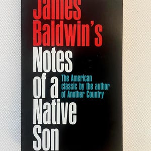 Könnte beinhalten: Ein Taschenbuch mit schwarzem Einband. Der Titel "James Baldwin's Notes of a Native Son" ist in Weiß und Rot dargestellt. Der Text "The American classic by the author of Another Country" ist ebenfalls sichtbar.