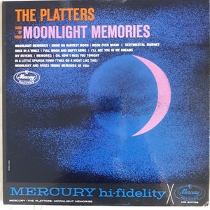 Peut inclure: Une pochette de disque vinyle vintage avec un croissant de lune rose sur un fond bleu. La pochette est pour l'album "Moonlight Memories" de "The Platters" sur Mercury Records. L'album comprend des chansons comme "Moonlight Memories", "Shine on Harvest Moon", "Moon Over Miami" et "Sentimental Journey".