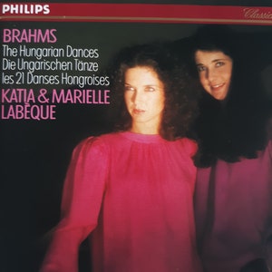 May include: A vintage album cover featuring a photo of two women in pink and purple shirts. The album title is "BRAHMS The Hungarian Dances Die Ungarischen Tänze les 21 Danses Hongroises KATIA & MARIELLE LABEQUE" and the record label is "PHILIPS Classics".