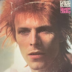 May include: Album cover featuring David Bowie with orange hair and a close-up of his face. The album title "Space Oddity" is in pink and the artist's name "David Bowie" is in white. The album cover has a vintage, slightly worn appearance.