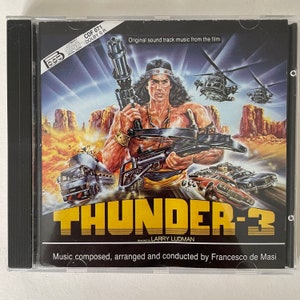 Puede incluir: Una caja de CD con una imagen colorida de un hombre sosteniendo un arma grande. El texto en la caja dice "Thunder-3" y "Música compuesta, arreglada y dirigida por Francesco de Masi".