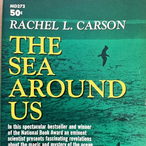 May include: A vintage paperback book cover with a yellow title "THE SEA AROUND US" by Rachel L. Carson. The cover features a green and blue ocean scene with a silhouette of a bird flying over the water.