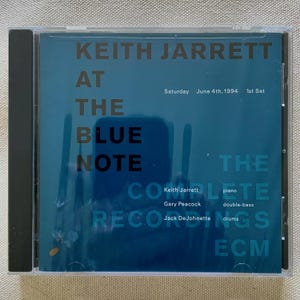 May include: A square CD jewel case featuring a dark teal cover with black text reading Keith Jarrett At The Blue Note The Complete Recordings. The cover includes the date Saturday June 4th, 1994 1st Set and lists musicians Keith Jarrett, Gary Peacock, and Jack DeJohnette with their respective instruments: piano, double-bass, and drums, plus the ECM logo.