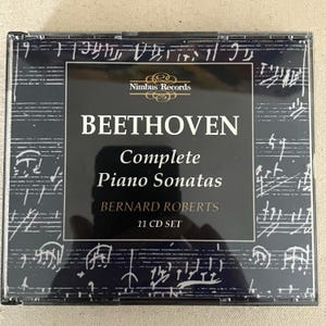 May include: A black and white CD box set titled Beethoven Complete Piano Sonatas by Bernard Roberts. The cover features a musical score background with white text on a black central panel, indicating an 11 CD set from Nimbus Records.
