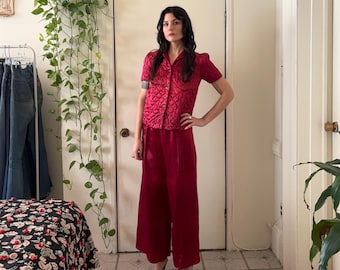 Puede incluir: Un conjunto burdeos de dos piezas con una camisa de manga corta con botones y un patrón texturizado y pantalones de pernera ancha. El conjunto se combina con sandalias de tacón negras y esponjosas. El fondo incluye una puerta blanca y plantas verdes.