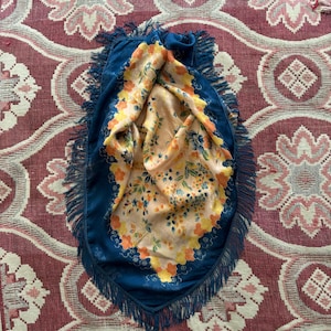 Puede incluir: Un pa&ntilde;uelo azul con flecos y un interior con estampado floral. El interior del pa&ntilde;uelo presenta un fondo melocot&oacute;n con flores naranjas, amarillas y azules. El pa&ntilde;uelo est&aacute; sobre una superficie roja y blanca estampada.