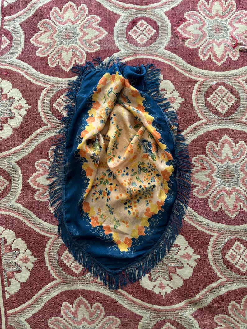 Puede incluir: Un pa&ntilde;uelo azul con flecos y un interior con estampado floral. El interior del pa&ntilde;uelo presenta un fondo melocot&oacute;n con flores naranjas, amarillas y azules. El pa&ntilde;uelo est&aacute; sobre una superficie roja y blanca estampada.