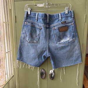 May include: Vintage blue denim Wrangler shorts with a distressed look. The shorts have a classic five-pocket design, a brown leather patch, and frayed edges. The shorts are hanging on a clear plastic hanger.