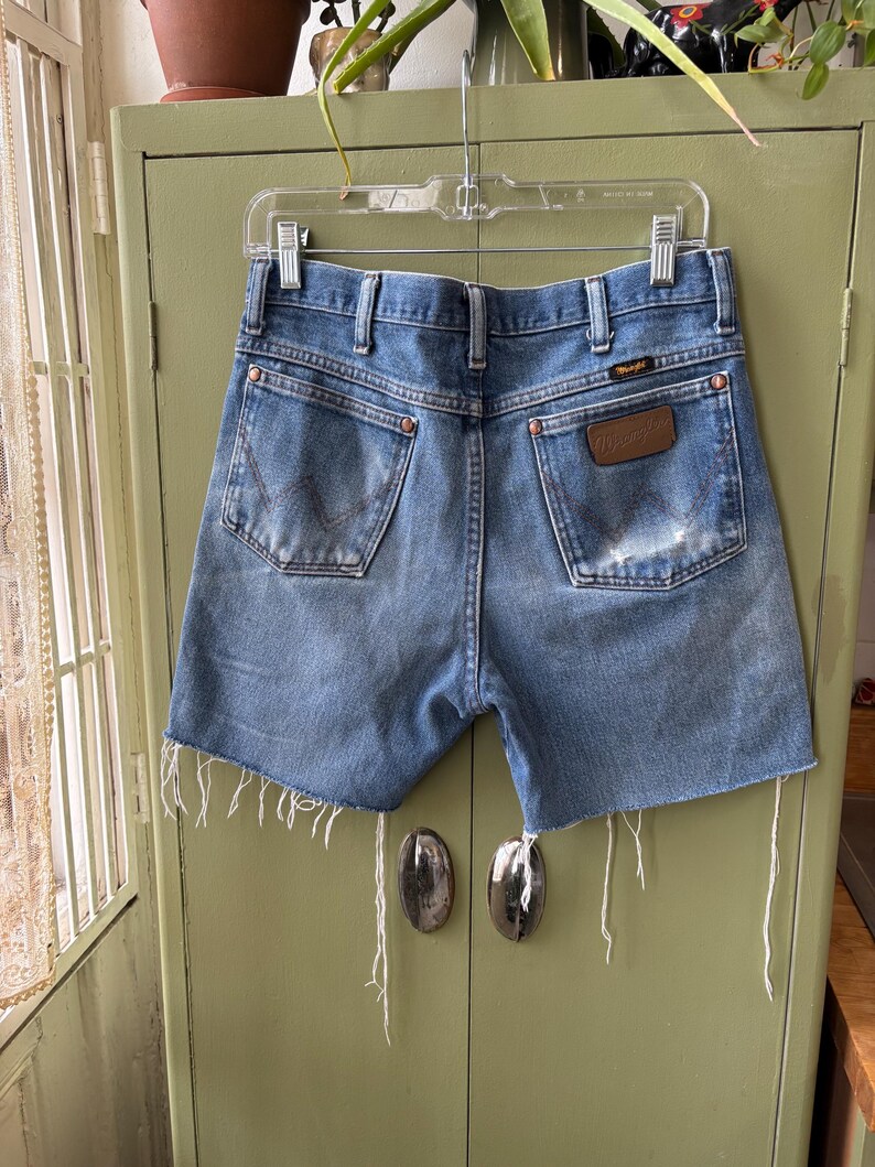 May include: Vintage blue denim Wrangler shorts with a distressed look. The shorts have a classic five-pocket design, a brown leather patch, and frayed edges. The shorts are hanging on a clear plastic hanger.