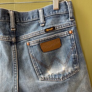 May include: Close-up of light blue denim Wrangler jeans. The back pocket features the Wrangler logo on a brown leather patch and a smaller logo above the pocket. Copper rivets and stitching detail the jeans.