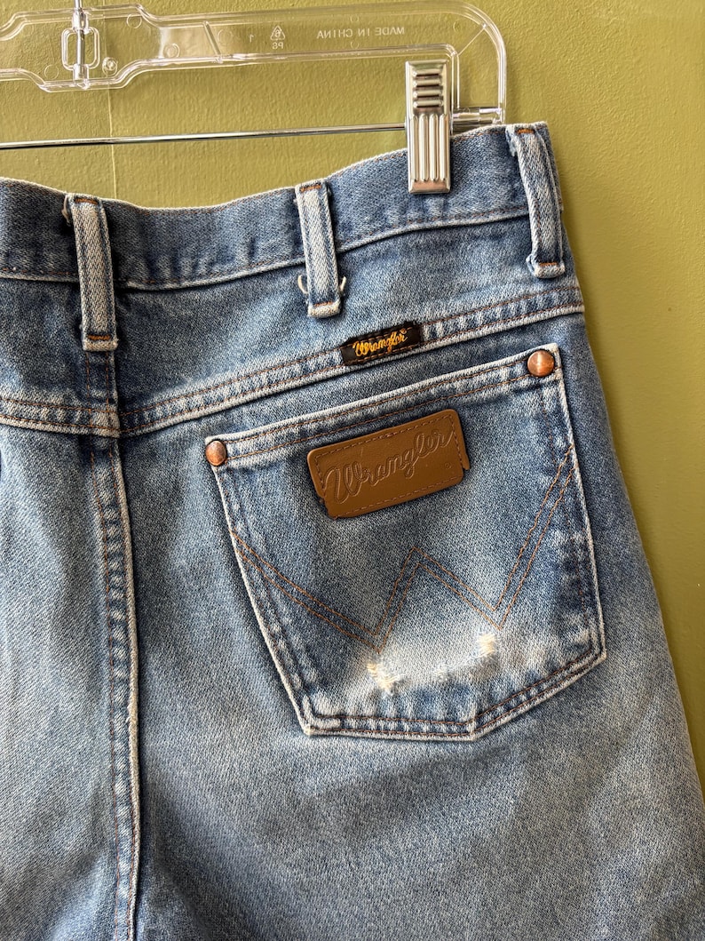 May include: Close-up of light blue denim Wrangler jeans. The back pocket features the Wrangler logo on a brown leather patch and a smaller logo above the pocket. Copper rivets and stitching detail the jeans.