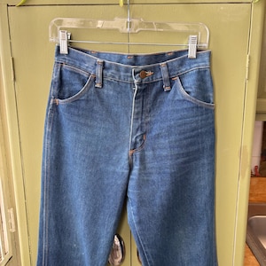 Puede incluir: Vaqueros azules de mezclilla con cierre de bot&oacute;n y dise&ntilde;o cl&aacute;sico de cinco bolsillos. Los vaqueros est&aacute;n colgados de una percha de pl&aacute;stico transparente. Las costuras son de color naranja en contraste.