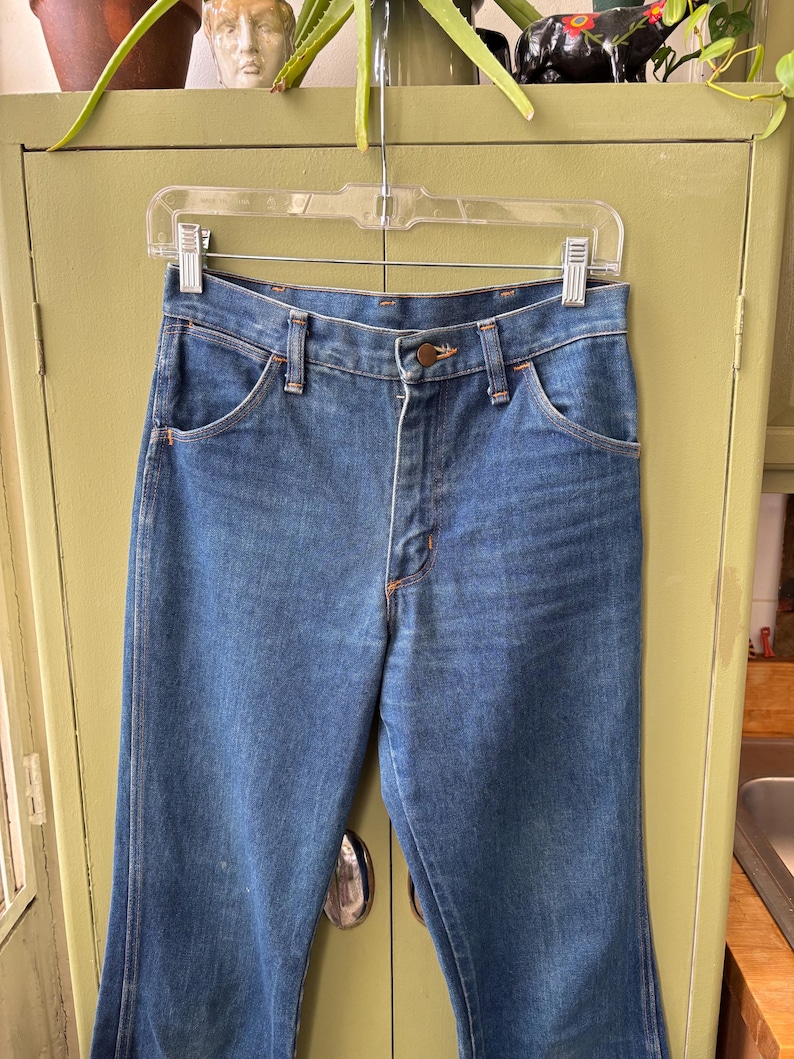 Puede incluir: Vaqueros azules de mezclilla con cierre de bot&oacute;n y dise&ntilde;o cl&aacute;sico de cinco bolsillos. Los vaqueros est&aacute;n colgados de una percha de pl&aacute;stico transparente. Las costuras son de color naranja en contraste.