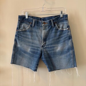 May include: Light blue denim shorts with a raw, frayed hem. The shorts have a classic five-pocket design and a button closure. The denim has a faded, worn appearance, suggesting a vintage style. The shorts are hanging on a clear plastic hanger.