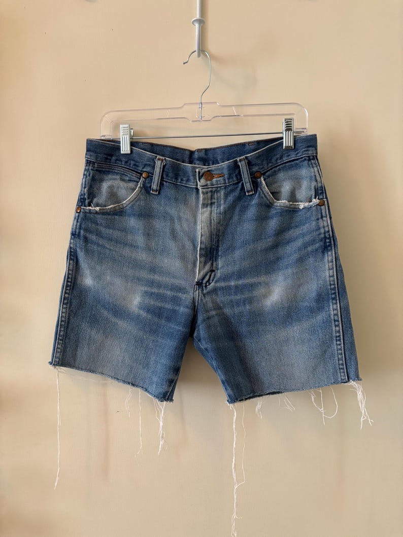 May include: Light blue denim shorts with a raw, frayed hem. The shorts have a classic five-pocket design and a button closure. The denim has a faded, worn appearance, suggesting a vintage style. The shorts are hanging on a clear plastic hanger.