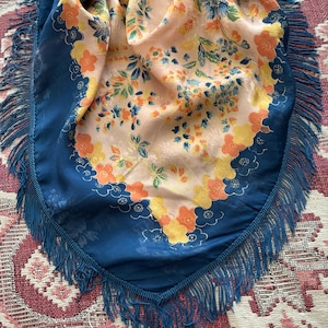 Puede incluir: Un pa&ntilde;uelo de seda vintage con un estampado floral en tonos naranja, amarillo y azul sobre un fondo melocot&oacute;n. El pa&ntilde;uelo tiene un borde azul oscuro y flecos.