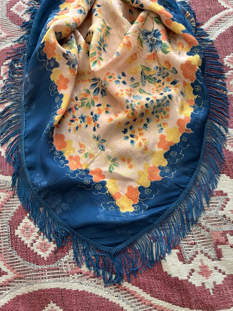 Puede incluir: Un pa&ntilde;uelo de seda vintage con un estampado floral en tonos naranja, amarillo y azul sobre un fondo melocot&oacute;n. El pa&ntilde;uelo tiene un borde azul oscuro y flecos.