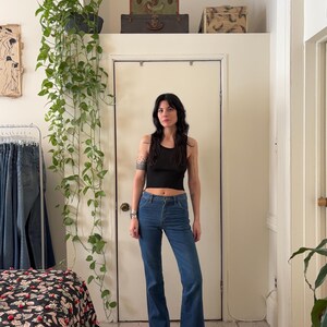 Puede incluir: Una persona con un top negro y vaqueros azules de campana. Los vaqueros son de cintura alta y se combinan con botas negras. El fondo incluye una puerta blanca, una planta colgante y maletas vintage.