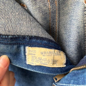 Puede incluir: Primer plano de unos vaqueros azules con una etiqueta visible. La etiqueta dice "WRANGLER NO FAULT DENIMS" e incluye la talla 29x32. Los vaqueros tienen una cremallera y est&aacute;n hechos de una tela vaquera duradera. Las costuras son visibles en hilo naranja.