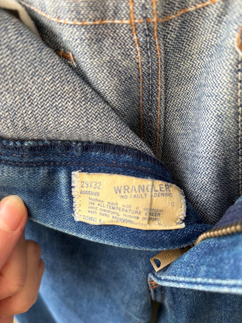 Puede incluir: Primer plano de unos vaqueros azules con una etiqueta visible. La etiqueta dice "WRANGLER NO FAULT DENIMS" e incluye la talla 29x32. Los vaqueros tienen una cremallera y est&aacute;n hechos de una tela vaquera duradera. Las costuras son visibles en hilo naranja.