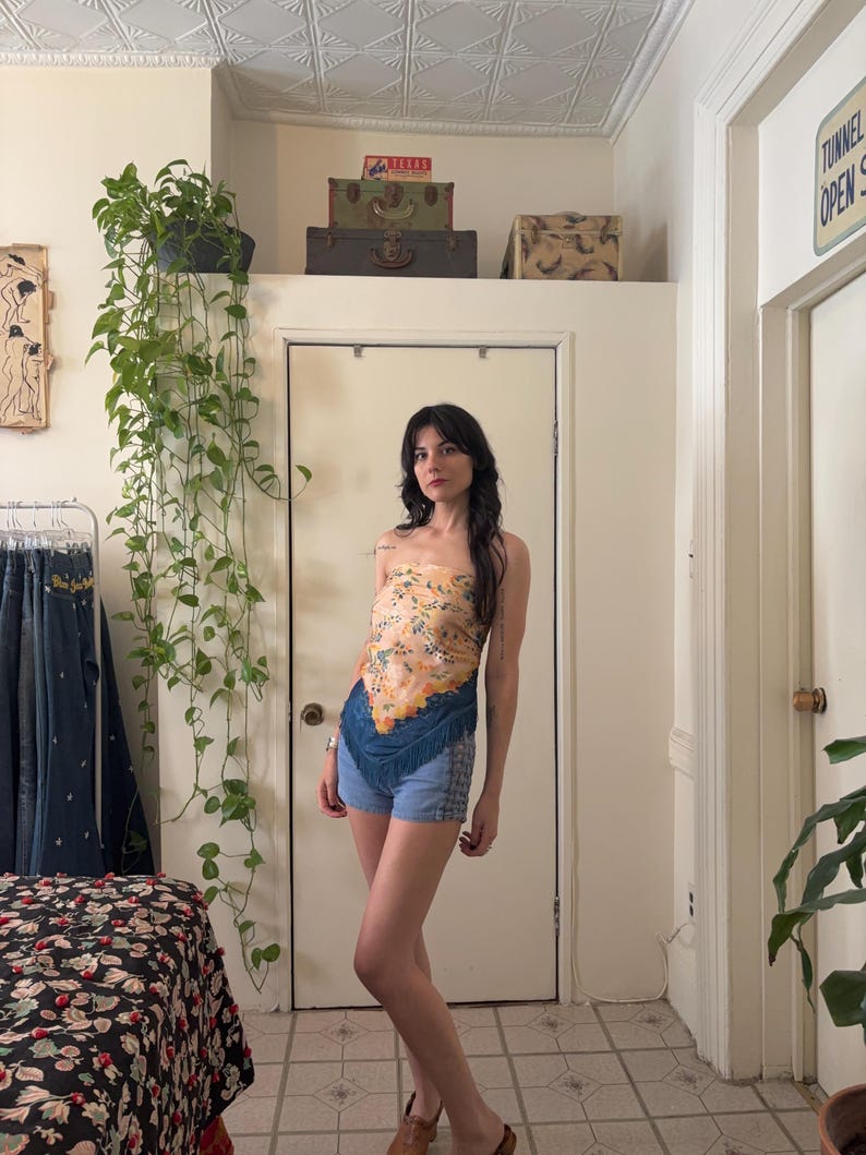Puede incluir: Una mujer con un top sin tirantes con estampado floral y pantalones cortos de mezclilla con flecos. El top es una mezcla de naranja, amarillo y azul. Los pantalones cortos son de mezclilla azul claro. Lleva sandalias marrones.