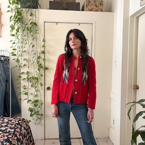 May include: A red knitted jacket with a southwestern-inspired design featuring embroidered stars and geometric patterns. The jacket has a button closure and two front pockets.