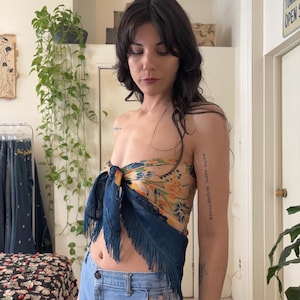 Puede incluir: Una mujer lleva un top de seda con un estampado floral en tonos amarillos, naranjas y azules. La bufanda est&aacute; anudada y tiene flecos azul oscuro. Lleva unos shorts vaqueros azul claro.