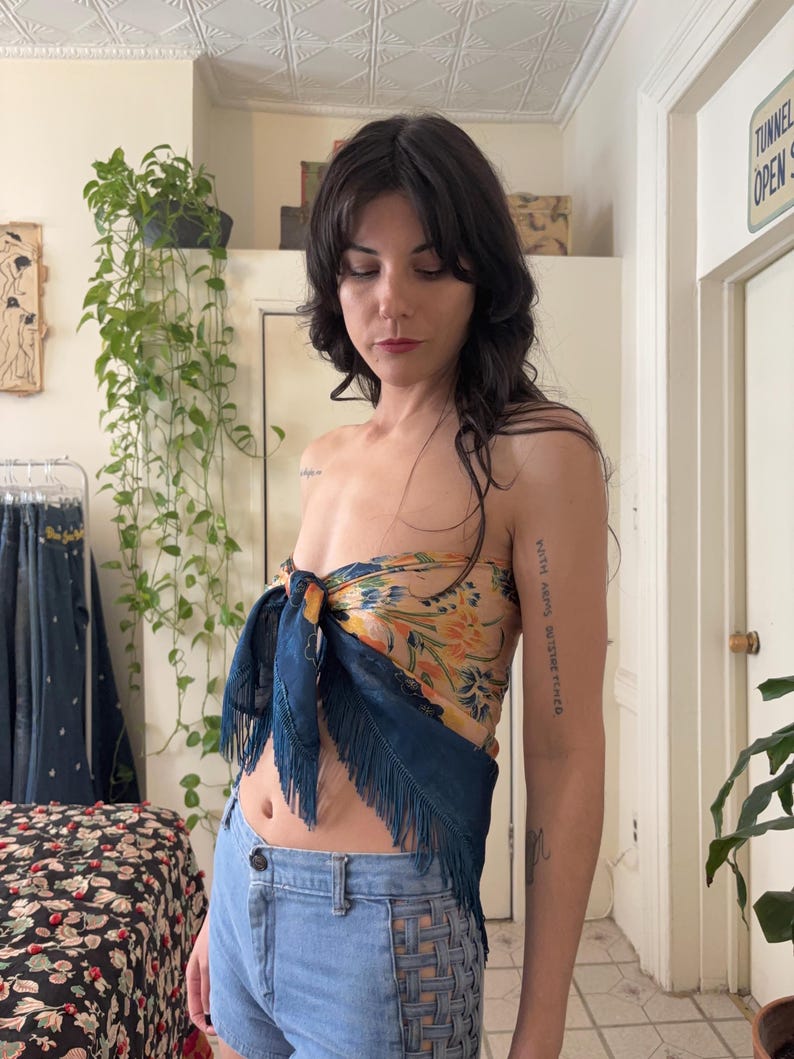 Puede incluir: Una mujer lleva un top de seda con un estampado floral en tonos amarillos, naranjas y azules. La bufanda est&aacute; anudada y tiene flecos azul oscuro. Lleva unos shorts vaqueros azul claro.