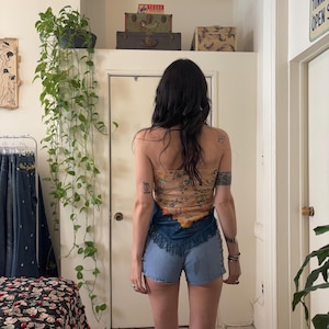 Puede incluir: Una mujer con un top sin tirantes con estampado floral y pantalones cortos de mezclilla con flecos. El top es de color beige claro con dise&ntilde;os florales naranjas y amarillos. Los pantalones cortos de mezclilla son de color azul claro.
