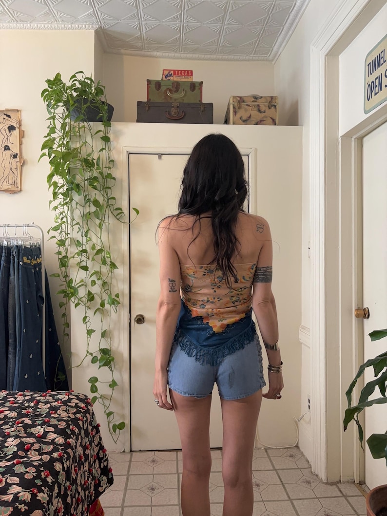 Puede incluir: Una mujer con un top sin tirantes con estampado floral y pantalones cortos de mezclilla con flecos. El top es de color beige claro con dise&ntilde;os florales naranjas y amarillos. Los pantalones cortos de mezclilla son de color azul claro.