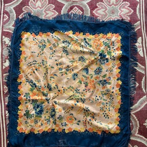 Puede incluir: Un pa&ntilde;uelo cuadrado con un estampado floral en tonos melocot&oacute;n, azul, naranja y amarillo. El pa&ntilde;uelo tiene un borde azul oscuro con flecos. El pa&ntilde;uelo se muestra sobre una superficie estampada roja y blanca.