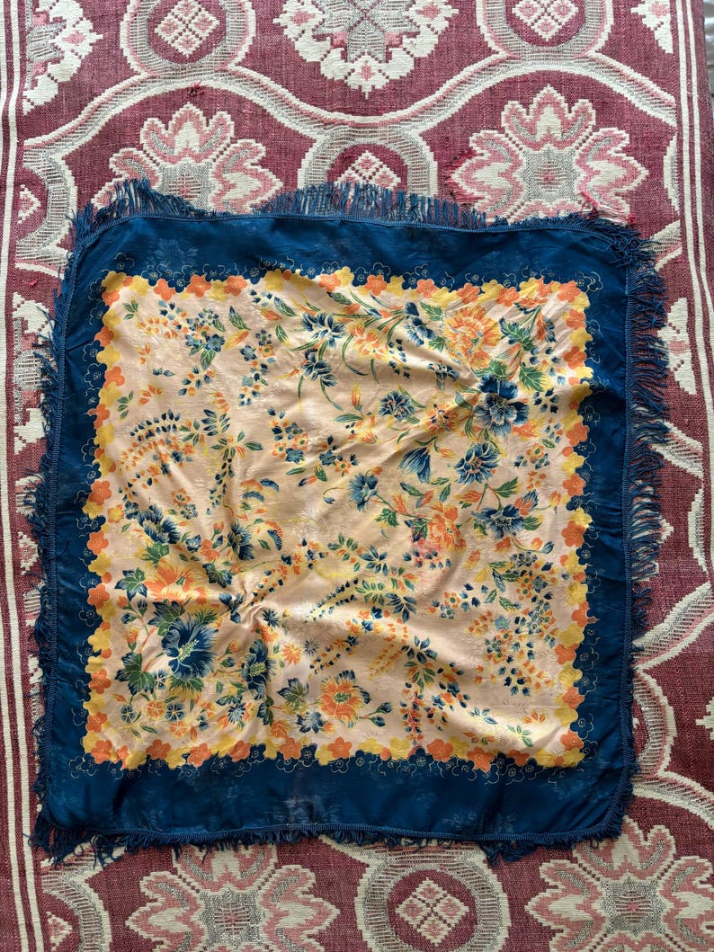 Puede incluir: Un pa&ntilde;uelo cuadrado con un estampado floral en tonos melocot&oacute;n, azul, naranja y amarillo. El pa&ntilde;uelo tiene un borde azul oscuro con flecos. El pa&ntilde;uelo se muestra sobre una superficie estampada roja y blanca.