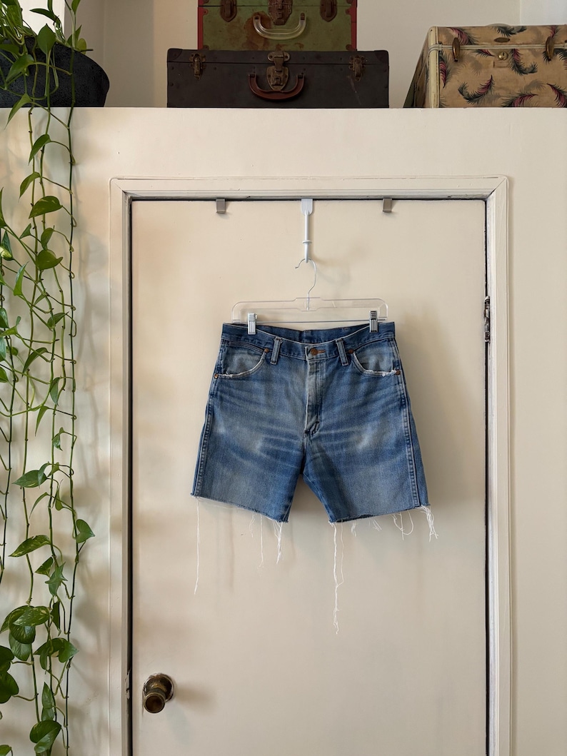 May include: Blue denim shorts with a raw hem, hanging on a white door. The shorts have a classic five-pocket design and a faded wash. The shorts are hanging on a metal hanger.