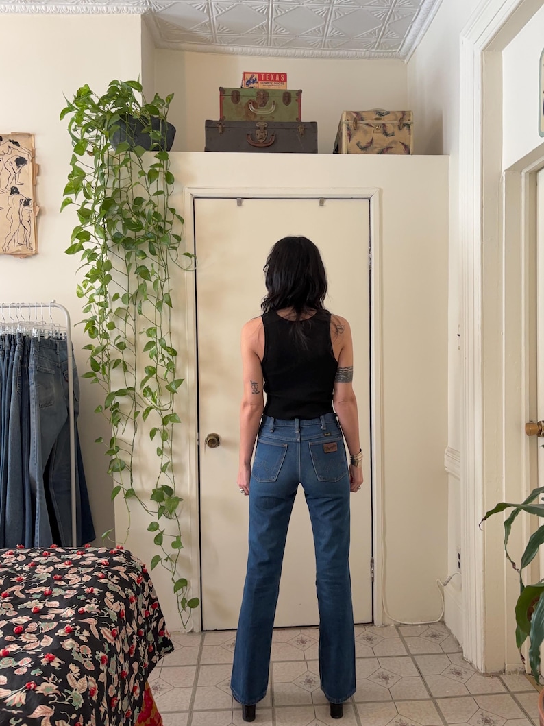 Puede incluir: Una persona lleva una camiseta sin mangas negra y unos vaqueros azules. Los vaqueros tienen un parche marr&oacute;n en el bolsillo trasero. La persona est&aacute; de pie frente a una puerta blanca. Hay plantas y maletas vintage en el fondo.