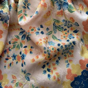 Puede incluir: Primer plano de una tela floral con fondo crema. La tela presenta un patr&oacute;n repetitivo de flores en tonos azules, naranjas y amarillos. Los bordes de la tela est&aacute;n ribeteados con un borde azul oscuro con flores naranjas y amarillas.