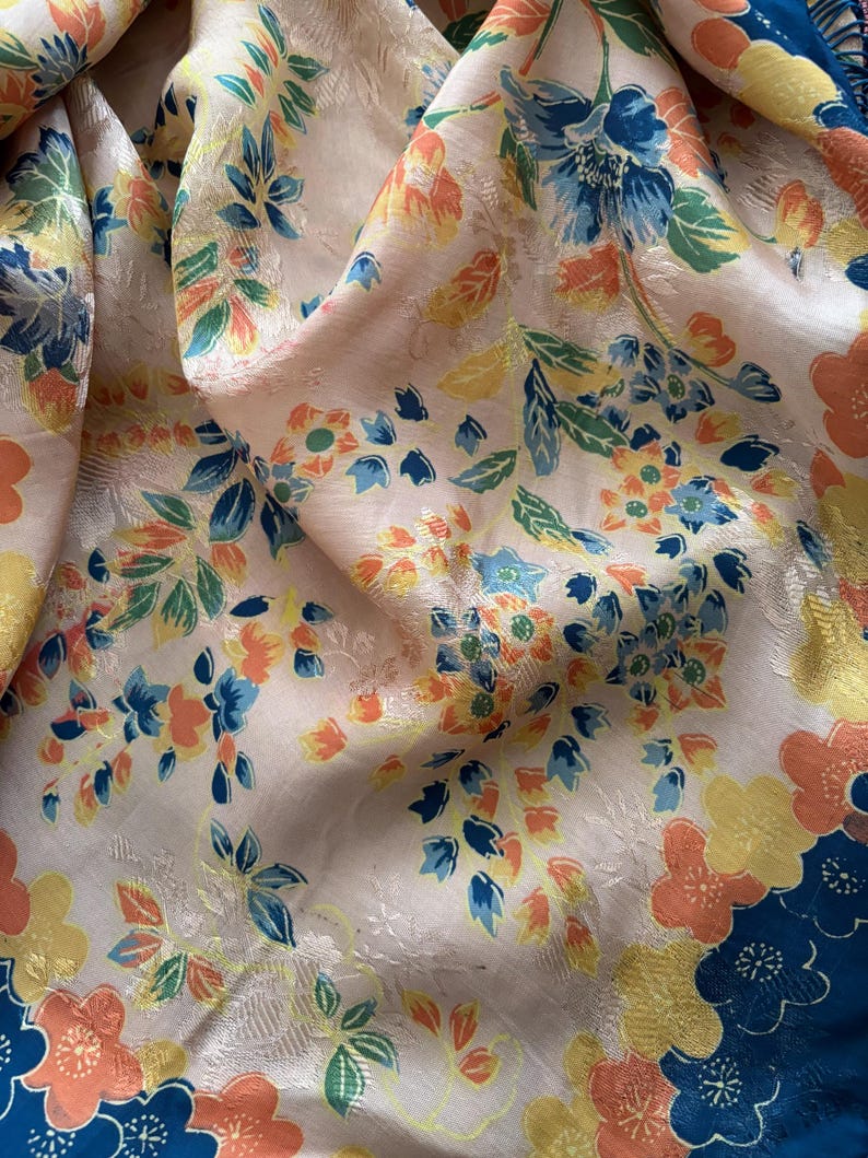 Puede incluir: Primer plano de una tela floral con fondo crema. La tela presenta un patr&oacute;n repetitivo de flores en tonos azules, naranjas y amarillos. Los bordes de la tela est&aacute;n ribeteados con un borde azul oscuro con flores naranjas y amarillas.
