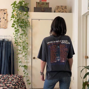 May include: A person wearing a dark gray Backstreet Boys concert t-shirt and blue jeans. The t-shirt features the band's logo and a list of tour dates. The person is standing in a room with a white door and a hanging plant.