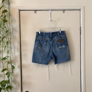 May include: Light blue denim shorts with a distressed look, hanging on a clear plastic hanger. The shorts have a brown leather patch on the back pocket and frayed edges. The shorts are hanging on a white door.