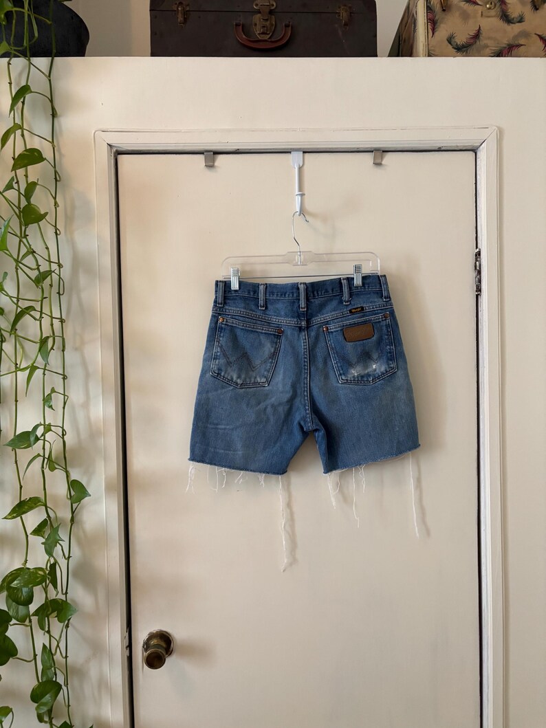 May include: Light blue denim shorts with a distressed look, hanging on a clear plastic hanger. The shorts have a brown leather patch on the back pocket and frayed edges. The shorts are hanging on a white door.