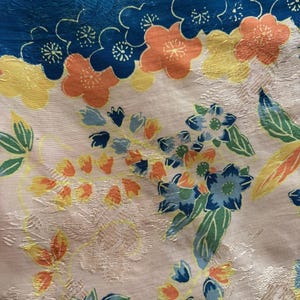 Puede incluir: Primer plano de una tela floral con un borde azul. La tela presenta un estampado de flores en tonos naranja, amarillo y azul, con hojas verdes sobre un fondo crema. La tela tiene un aspecto texturizado.