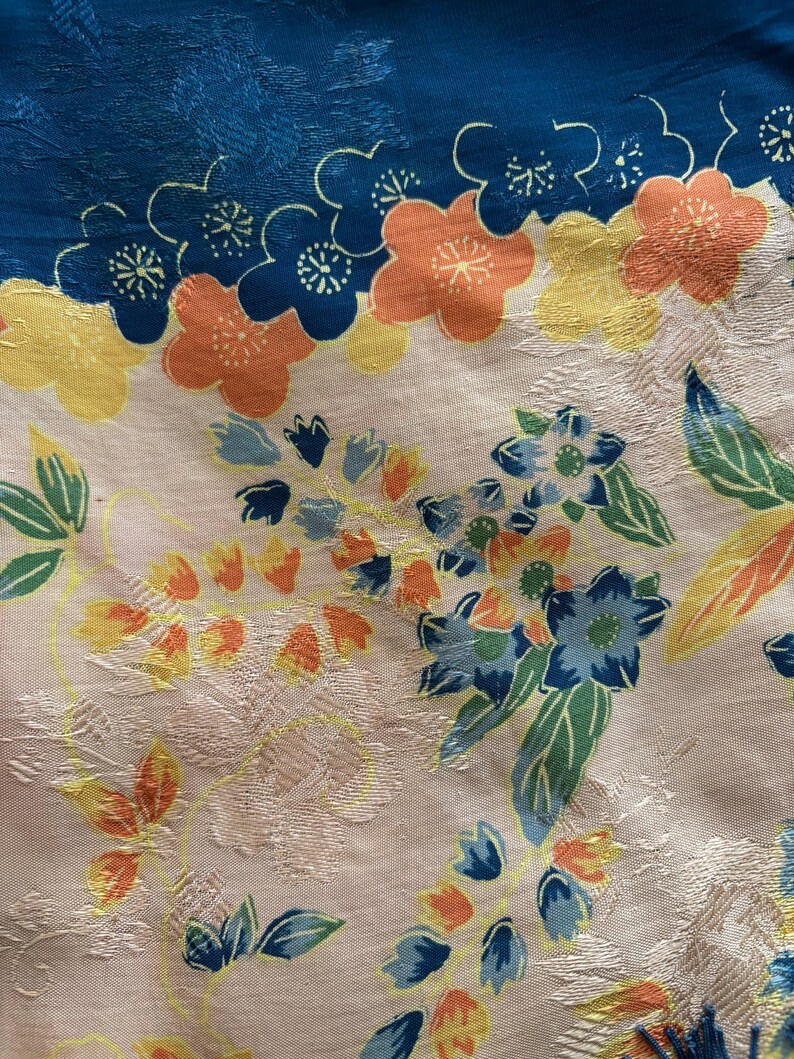 Puede incluir: Primer plano de una tela floral con un borde azul. La tela presenta un estampado de flores en tonos naranja, amarillo y azul, con hojas verdes sobre un fondo crema. La tela tiene un aspecto texturizado.