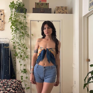 Puede incluir: Una mujer con un top sin tirantes con estampado floral y detalles de plumas azules, combinado con shorts vaqueros azul claro. El top est&aacute; atado con un lazo. El fondo incluye una habitaci&oacute;n con plantas y maletas vintage.