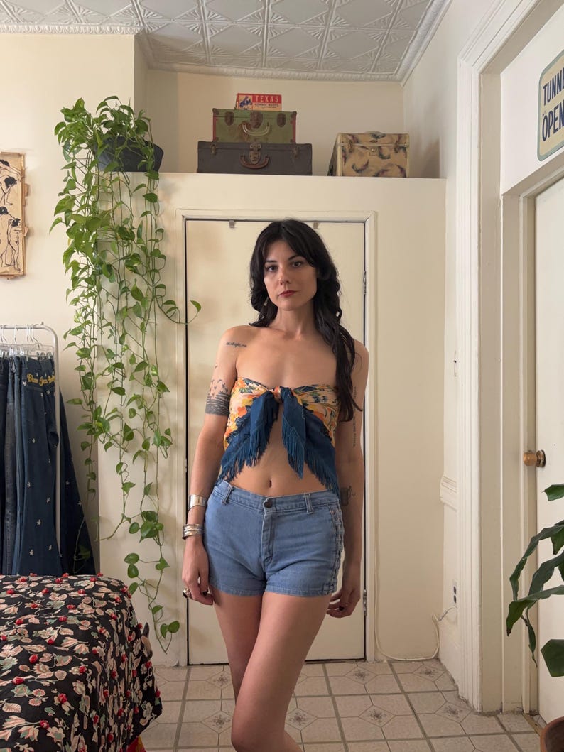 Puede incluir: Una mujer con un top sin tirantes con estampado floral y detalles de plumas azules, combinado con shorts vaqueros azul claro. El top est&aacute; atado con un lazo. El fondo incluye una habitaci&oacute;n con plantas y maletas vintage.