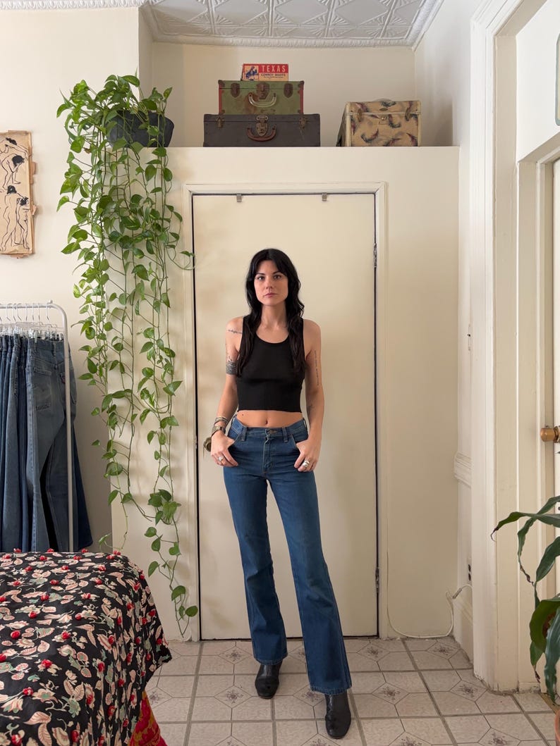 Puede incluir: Una persona lleva un top corto negro y vaqueros azules con pernera acampanada. El fondo incluye una puerta blanca, una cama con una colcha estampada floral y plantas colgantes.