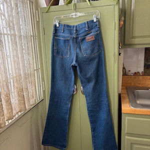 Puede incluir: Vaqueros Wrangler de mezclilla azul colgados de una percha transparente. Los vaqueros tienen un dise&ntilde;o cl&aacute;sico de cinco bolsillos con un parche de cuero marr&oacute;n en el bolsillo trasero. Los vaqueros son de un lavado medio con una ligera campana en la parte inferior.