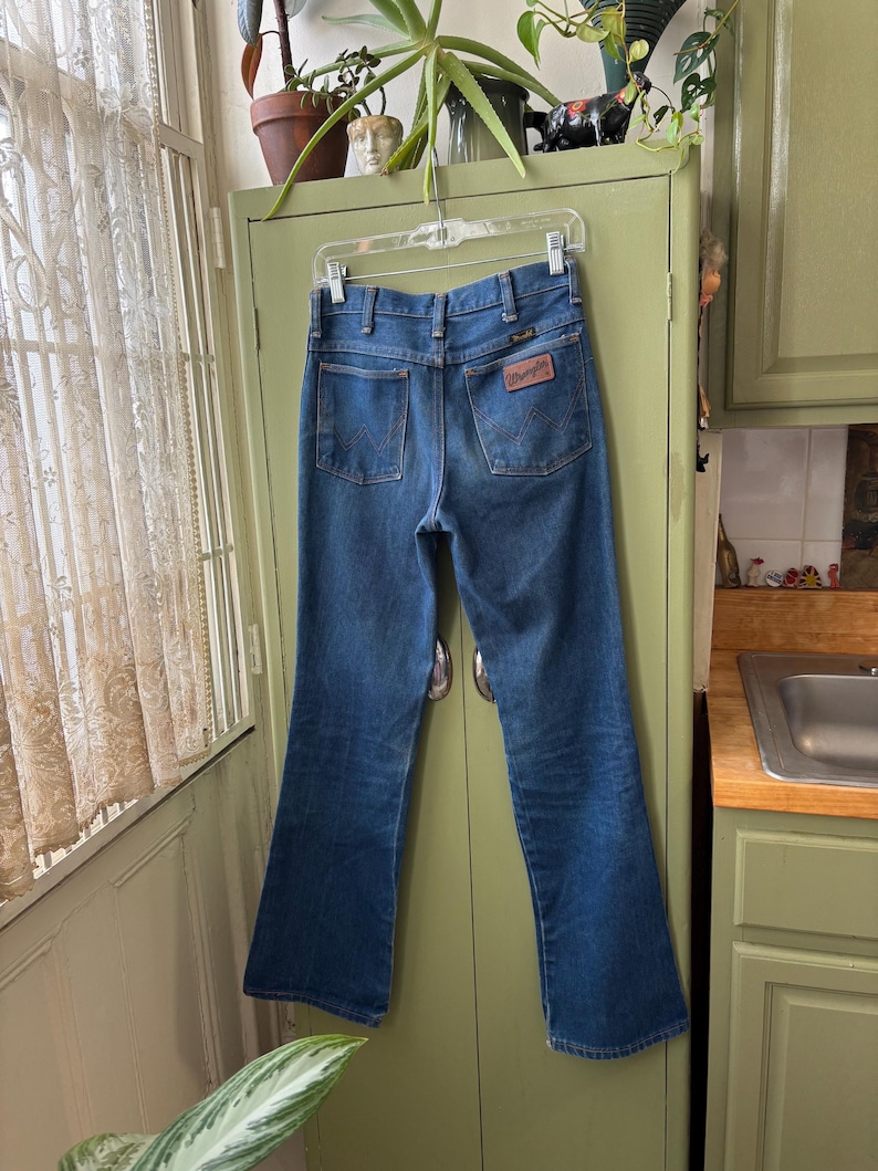 Puede incluir: Vaqueros Wrangler de mezclilla azul colgados de una percha transparente. Los vaqueros tienen un dise&ntilde;o cl&aacute;sico de cinco bolsillos con un parche de cuero marr&oacute;n en el bolsillo trasero. Los vaqueros son de un lavado medio con una ligera campana en la parte inferior.
