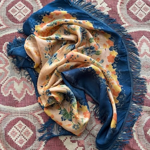 Puede incluir: Bufanda azul y melocot&oacute;n con estampado floral. La bufanda tiene un borde azul oscuro con flecos. El estampado floral incluye flores naranjas, amarillas y verdes. La bufanda est&aacute; hecha de una tela suave y fluida.