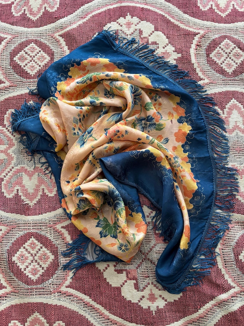 Puede incluir: Bufanda azul y melocot&oacute;n con estampado floral. La bufanda tiene un borde azul oscuro con flecos. El estampado floral incluye flores naranjas, amarillas y verdes. La bufanda est&aacute; hecha de una tela suave y fluida.