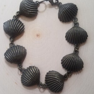 May include: A bronze bracelet with a chain and seven shell-shaped charms. The bracelet is on a light wood surface.